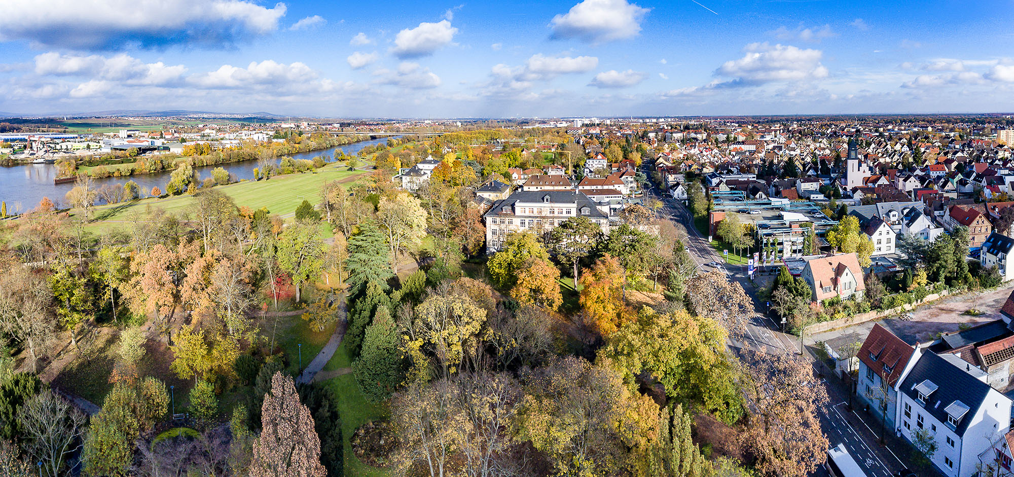 Luftaufnahme von Rüsselsheim am Main im Herbst mit dem weitläufigen Schlosspark, dem historischen Schloss und dem Main im Hintergrund.