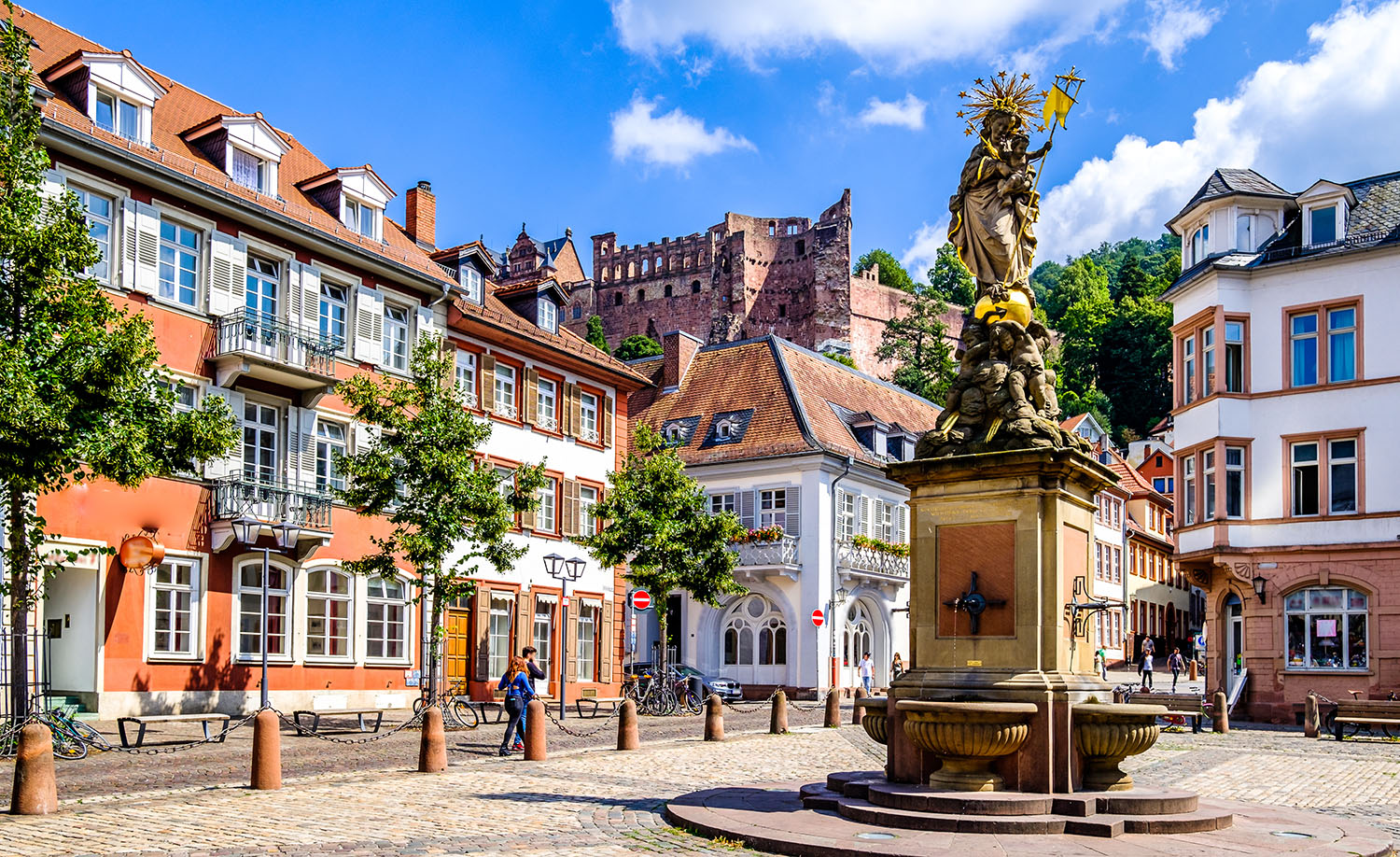 Kornmarkt in der Heidelberger Altstadt mit dem historischen Marienbrunnen, umgeben von charakteristischen Fachwerkfassaden und den Ruinen des Heidelberger Schlosses im Hintergrund.