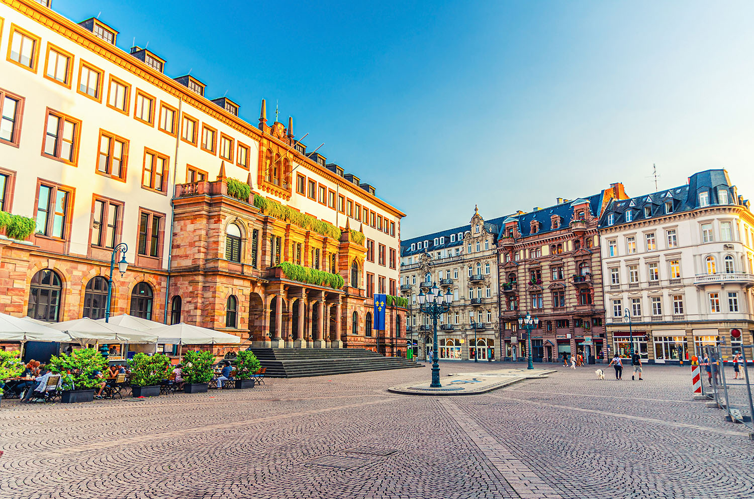 Blick auf den Wiesbadener Marktplatz mit dem historischen Rathaus aus rotem Sandstein, eleganten Gründerzeitgebäuden und einem belebten Straßencafé im Vordergrund.