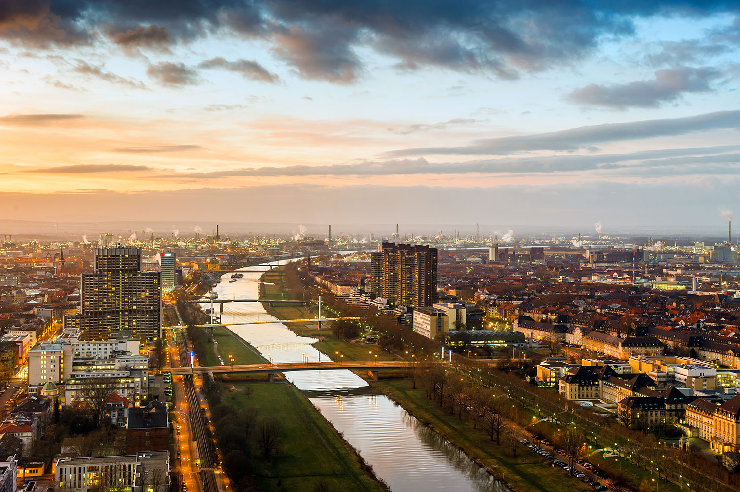 Abendliche Luftaufnahme von Mannheim mit dem beleuchteten Neckar, der Stadtsilhouette und dem Industriegebiet von Ludwigshafen im Hintergrund bei Sonnenuntergang.