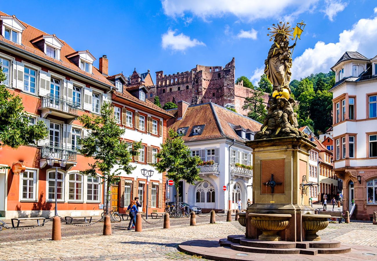 Kornmarkt in der Heidelberger Altstadt mit dem historischen Marienbrunnen, umgeben von charakteristischen Fachwerkfassaden und den Ruinen des Heidelberger Schlosses im Hintergrund.