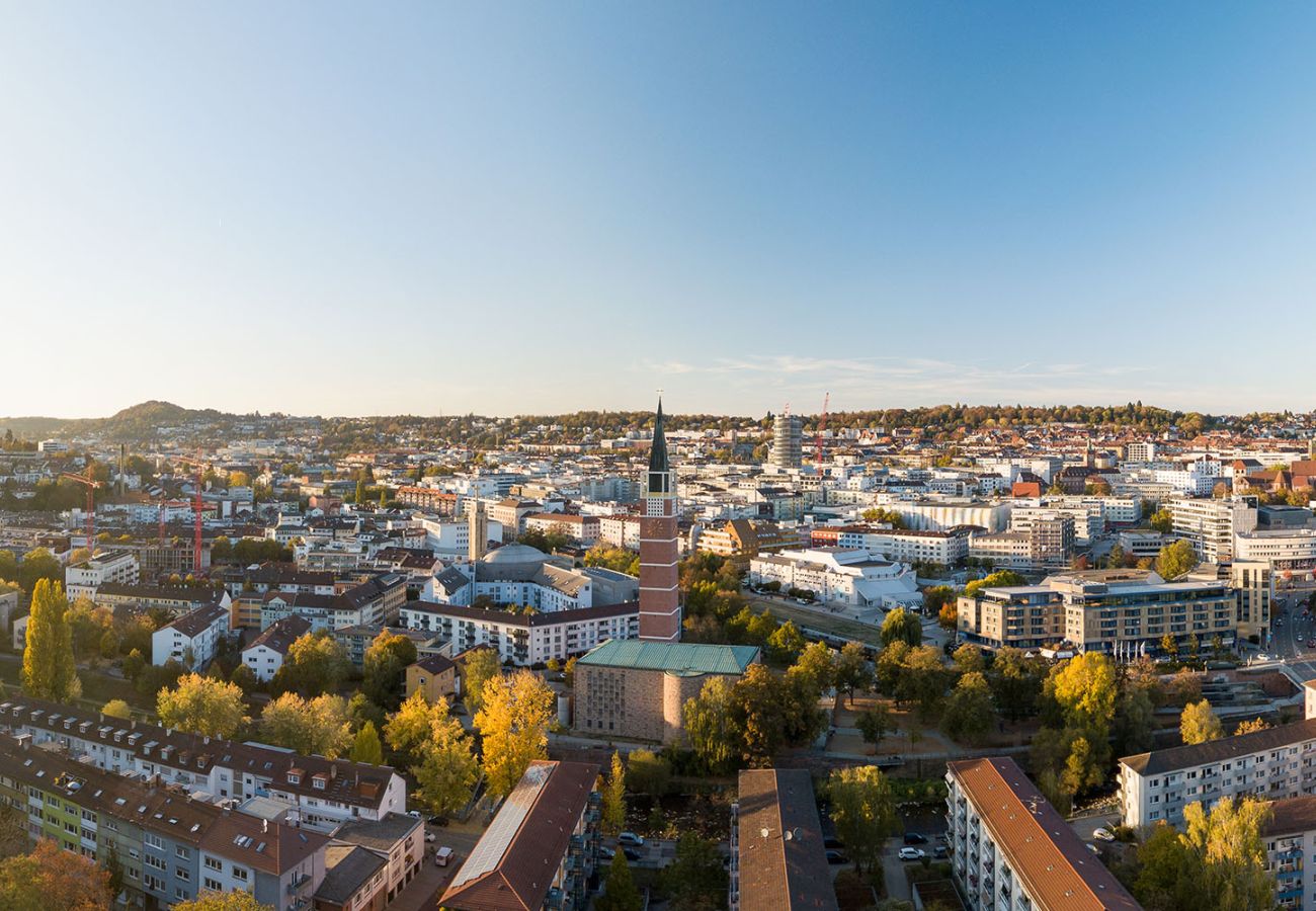 Luftaufnahme der Stadt Pforzheim im Herbst mit dem markanten Kirchturm der Stadtkirche im Zentrum, umgeben von buntem Laubwerk und der weitläufigen Stadtlandschaft Baden-Württembergs.