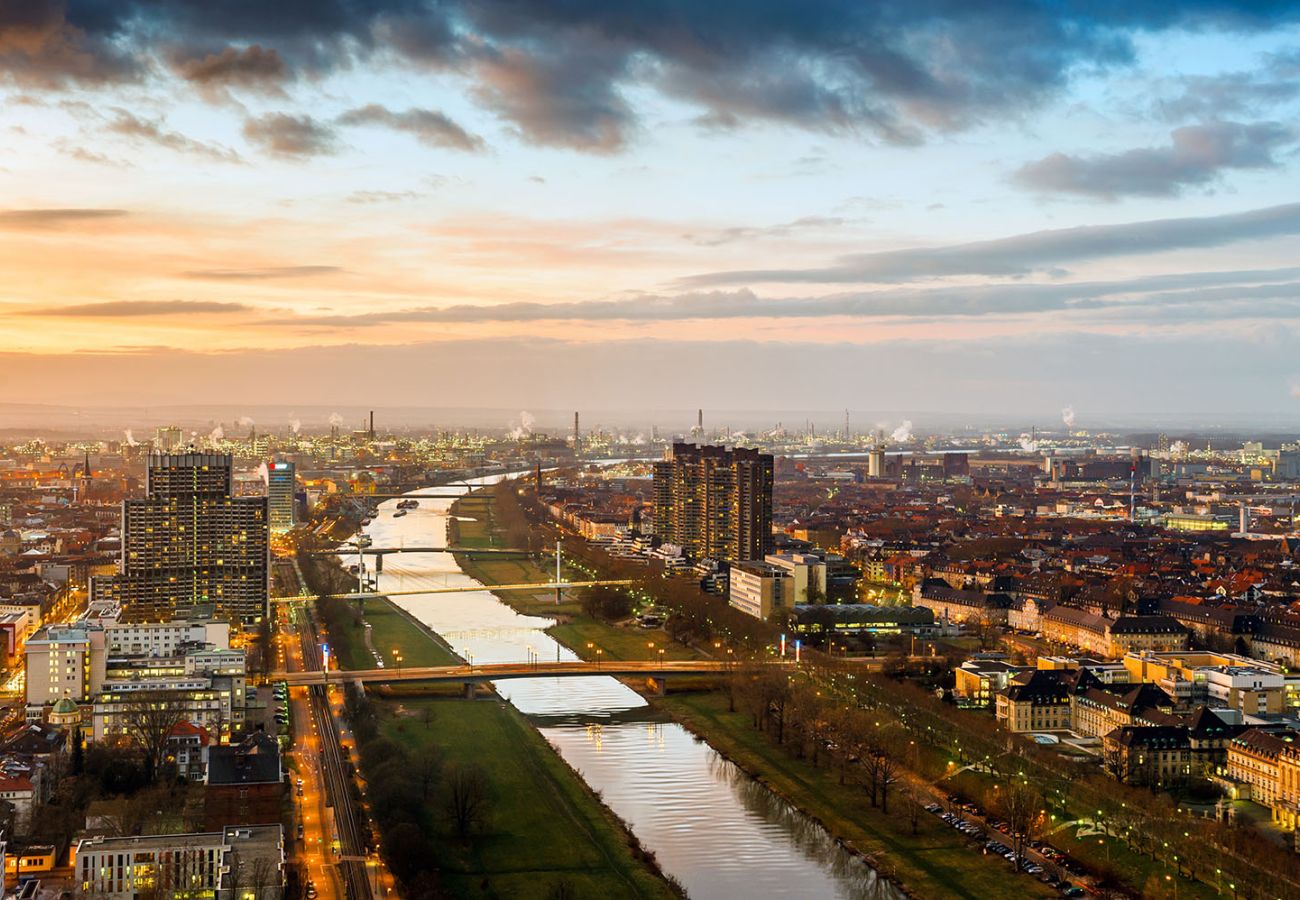 Abendliche Luftaufnahme von Mannheim mit dem beleuchteten Neckar, der Stadtsilhouette und dem Industriegebiet von Ludwigshafen im Hintergrund bei Sonnenuntergang.
