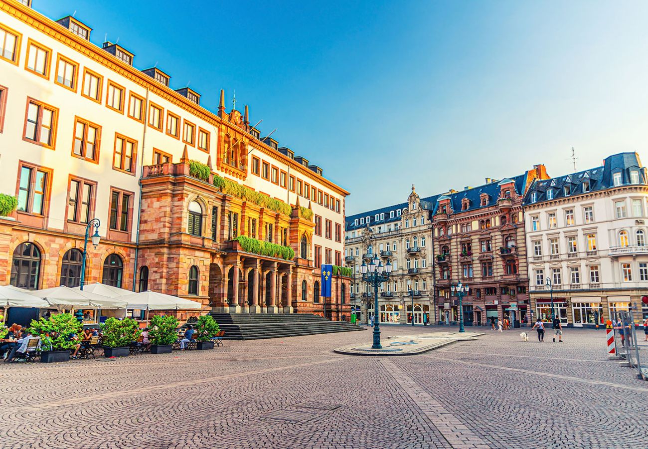 Blick auf den Wiesbadener Marktplatz mit dem historischen Rathaus aus rotem Sandstein, eleganten Gründerzeitgebäuden und einem belebten Straßencafé im Vordergrund.